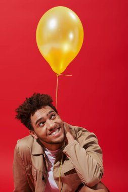 Kırmızı arka planda sarı balonun yanında gülümseyen bej ceketli Afro-Amerikan adam.