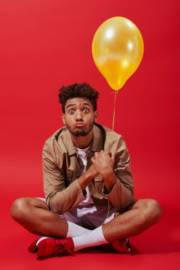 Kırmızı arka planda sarı balonla oturan bej ceketli komik, Afro-Amerikan bir adam.