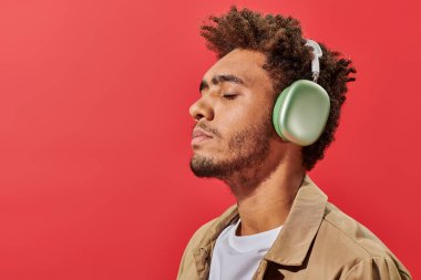 Kablosuz kulaklıklı genç Afrikalı Amerikalı adamın portresi kırmızı arka planda müzik dinliyor.
