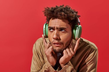 Kafası karışmış, kablosuz kulaklıklı, kırmızı arka planda müzik dinleyen Afro-Amerikan bir adamın portresi.