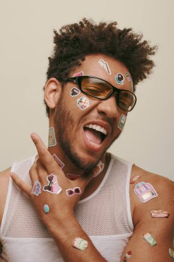 expressive african american fella in sunglasses with trendy stickers on face on grey background clipart