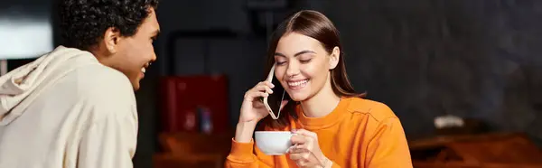 happy young woman chatting on her smartphone with a coffee cup near black boyfriend in cafe, banner
