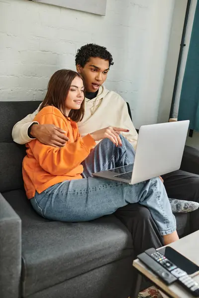 stock image diverse couple sitting on couch and browsing laptop, woman pointing at screen near black boyfriend