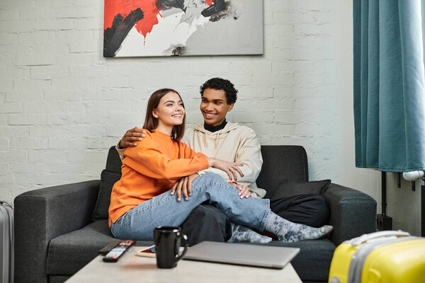 cheerful diverse couple enjoys cozy moment on a sofa in hostel, with travel bags indicating a trip