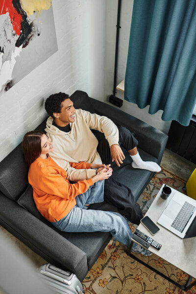 jolly diverse couple enjoys a calm moment while relaxing on sofa with a gadgets and luggage