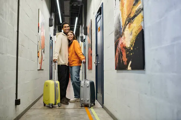 Relaxed multicultural couple with luggage smiling and standing together in a hostel hallway