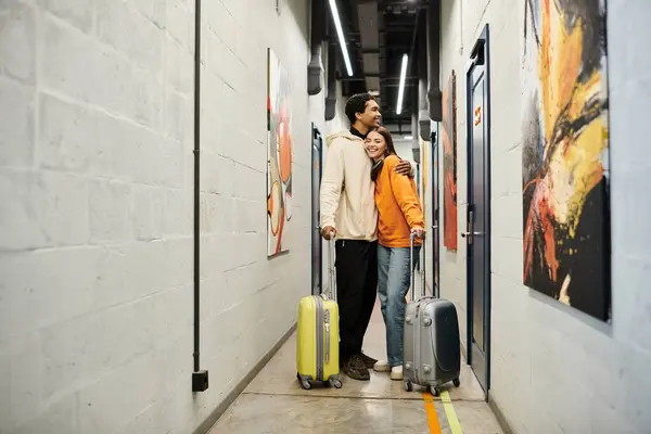 Relaxed multicultural couple with luggage embracing and standing together in a hostel hallway