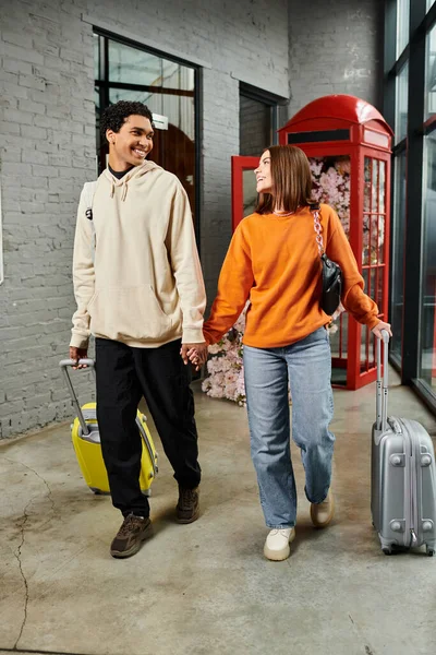 interracial smiling couple walks through a corridor holding hands and pulling a suitcase, travel