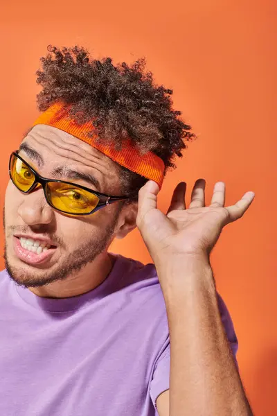 stock image young curious african american man in sunglasses gesturing while listening on orange background