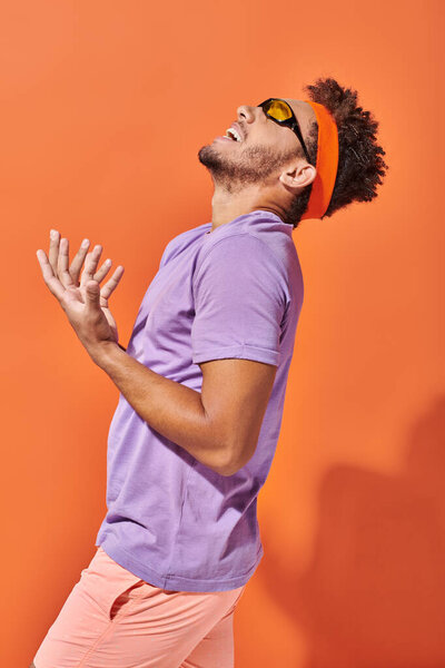 expressive african american man in eyeglasses and headband screaming on orange background, gesture
