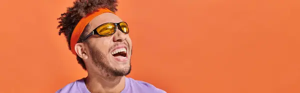 Stock image african american man in sunglasses and headband laughing and looking away on orange backdrop, banner