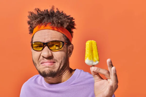stock image displeased african american man in sunglasses and headband looking at fruity ice cream on orange