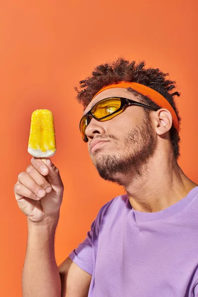stock image african american man in sunglasses and headband holding frozen ice cream on orange background