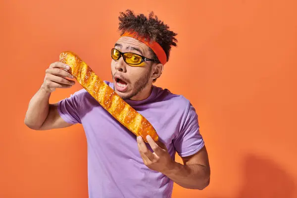 stock image african american man in sunglasses and headband biting fresh baguette on orange background, bakery