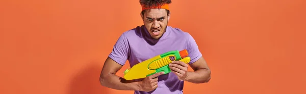 stock image african american man in headband playing water fight with toy gun on orange background, banner