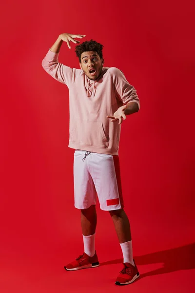 shocked african american man in gym clothes gesturing with opened mouth on red background