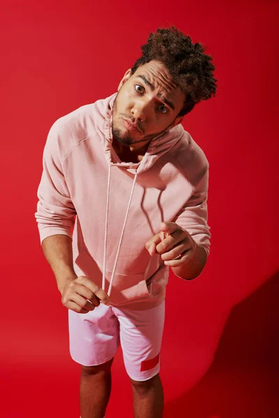 stock image african american man pulling drawstrings of pink hoodie and looking at camera on red background