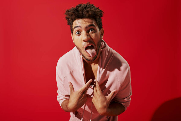 funny african american man pulling pink hoodie and sticking out tongue while on red background