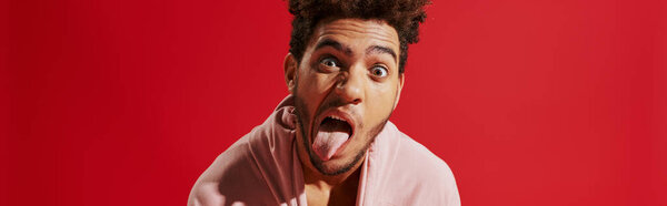 african american man pulling pink hoodie and sticking out tongue while on red background, banner