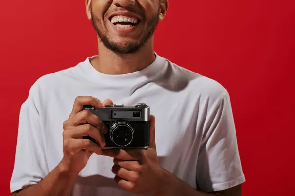 Stock image cropped and happy african american photographer holding vintage camera and smiling on red background