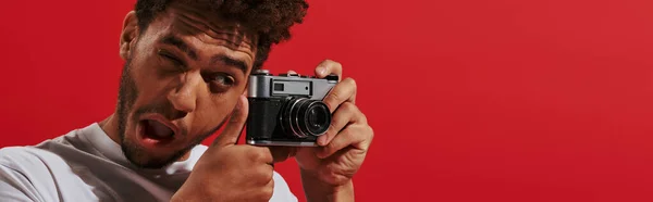 stock image funny african american man looking at viewfinder while taking shot on red background, banner