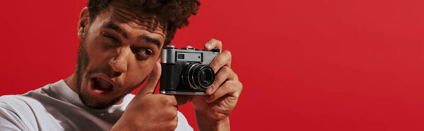 funny african american man looking at viewfinder while taking shot on red background, banner