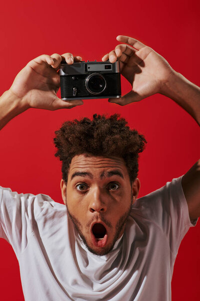 amazed african american photographer with open mouth taking shot on retro camera on red background