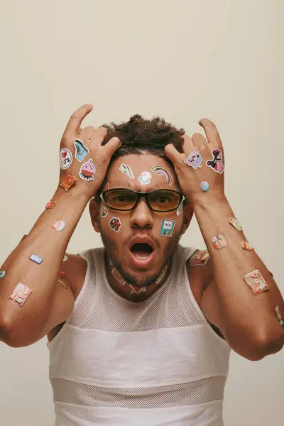 stock image shocked young african american man in sunglasses with trendy stickers on face on grey background