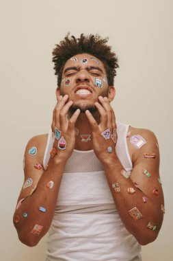 emotional african american man with stickers on face scratching beard on grey background, hipster