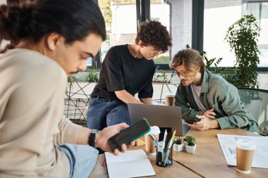 Genç profesyoneller telefonlu bulanık bir iş arkadaşının yanında, modern ofiste dizüstü bilgisayarla beyin fırtınası yapıyorlar.
