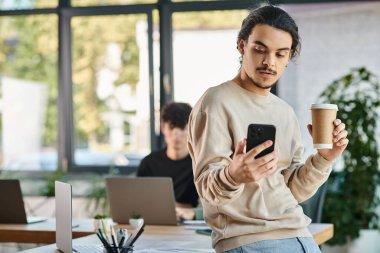 20 'li yaşlarda genç bir adam, akıllı telefon kullanıyor ve işe yeni başlayan bir ofiste kahve tutuyor.