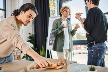 Kahveli genç adam rahatlamış ofis atmosferinde pizza yiyor, öğle yemeği molası veriyor.