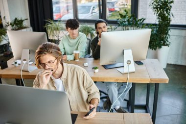 Yaratıcı proje üzerinde çalışan odaklanmış bir adam. Akıllı telefonu ofisinde tutarken monitöre bakıyor.