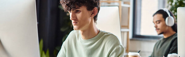 Young curly man engaged in retouching work on computer monitor near colleague in office, banner
