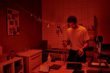 Siyah fotoğrafçının elindeki zamanlayıcı film işleme için geliştiriciyi tepsiye koyar, kırmızı karanlık oda.