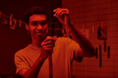 happy african american photographer examines developed film strip, bathed in darkroom with red light clipart