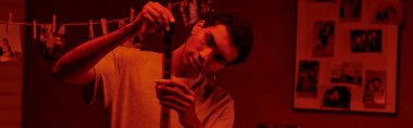 focused african american photographer examines developed film strip, bathed in red darkroom, banner clipart