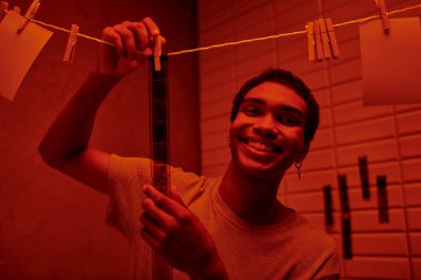 cheerful african american man hanging freshly developed film strip  in a red-lit darkroom, nostalgia clipart