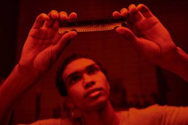 focused african american photographer examine developed film strip in a red-lit darkroom clipart