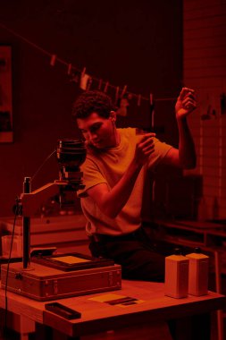 Genç Afrikalı Amerikalı fotoğrafçı, karanlık bir odanın kırmızı ışığı altında fotoğraf şeritlerini karşılaştırıyor.