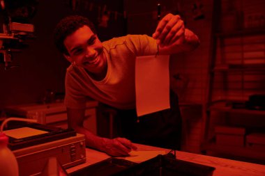 happy african american photographer holding tweezers with freshly developed photo paper in darkroom clipart