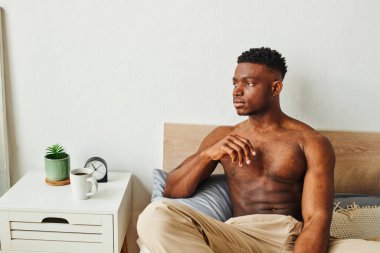 Genç, üstsüz, kaslı, Afro-Amerikalı bir adam bir fincan sabah kahvesiyle komodinin yanında oturuyor.