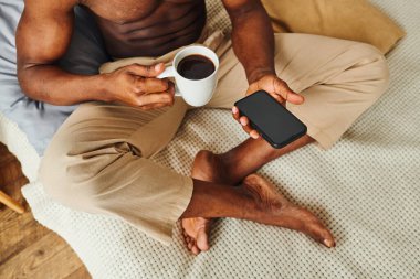 Afro-Amerikalı pijama pantolonlu bir adam kahve ve boş ekranlı bir akıllı telefonla yatakta oturuyor.