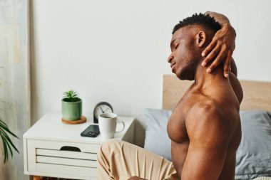 Spor tişörtsüz Afro-Amerikan bir adam. Yatak odasındaki sehpanın yanında boynu uzanıyor.
