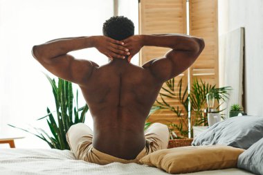 shirtless and muscular african american man sitting and stretching on bed in morning, back view clipart