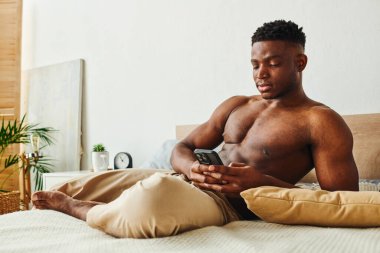 Atletik Afro-Amerikalı pijama pantolonlu adam sabah rahat bir yatakta cep telefonuyla mesajlaşıyor.