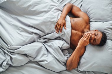 top view of sleepy african american man yawning while waking up on comfortable bed in morning clipart
