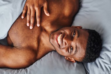 top view of young and muscular african american man with radiant smile looking at camera on bed clipart