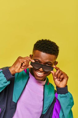 Havalı Afro-Amerikan adam şık güneş gözlüklü ve renkli ceketli sarı kameraya gülümsüyor.