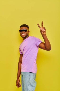 Güneş gözlüklü ve mor tişörtlü neşeli Afro-Amerikalı adam sarı üzerinde zafer işareti gösteriyor.